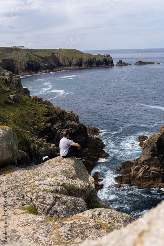 Obraz na płótnie Photo of a young and attractive man sitting on a rock next to a cliff in the middle of nature, in Land's End, UK. Amazing landscape in a sunny day. w salonie
