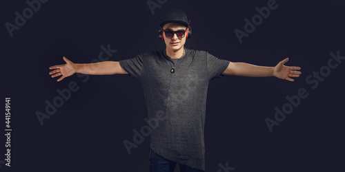 Portrait of attractive cheerful guy in sunglasses and baseball cap listening bass sound isolated over dark background