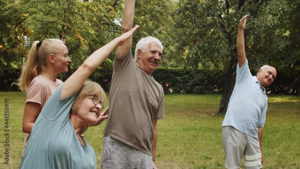 Young female trainer helping cheerful senior people with side stretch exercise during group fitness class outdoor in park