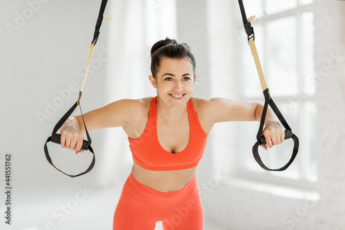 Athletic woman squeezing on suspension straps, doing trx exercise at white gym
