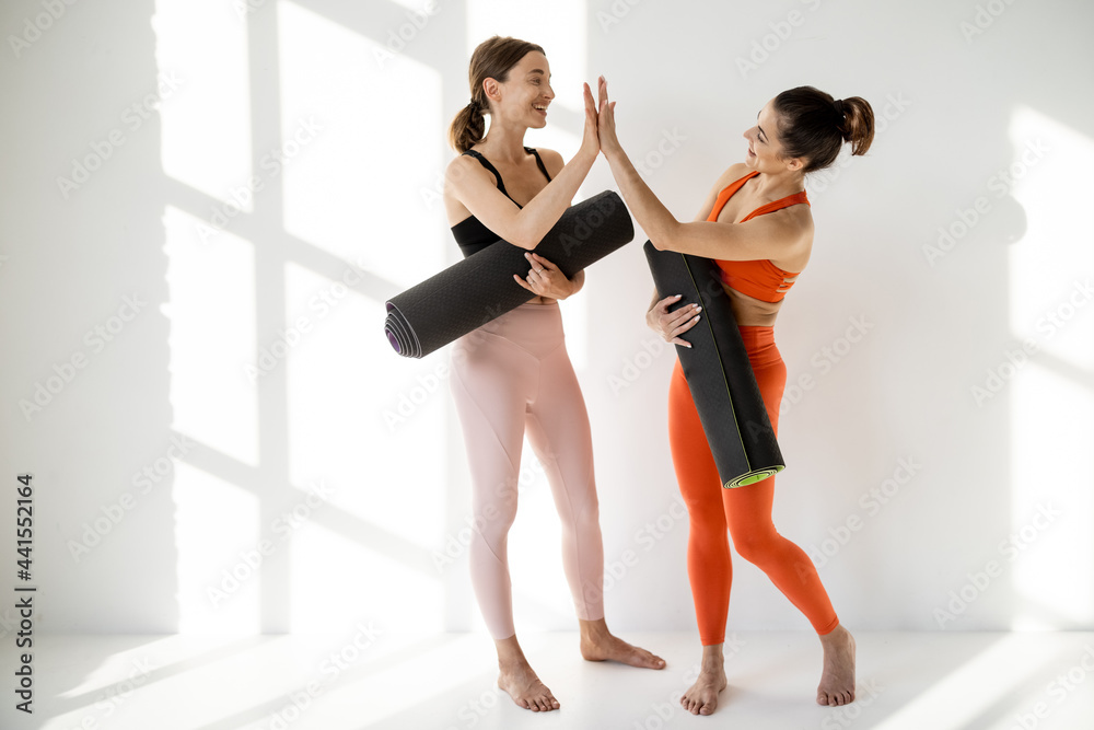 Obraz premium Two girlfriends greet each other before yoga training, standing on the white wall background with a window shadows