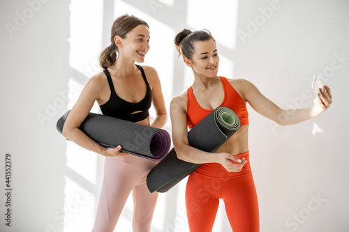 Fototapeta Naklejka Na Ścianę i Meble -  Two athletic sports women meet at gym, making selfie photo while standing together with yoga mats in the white hallway. Meeting friends and having fun on yoga training