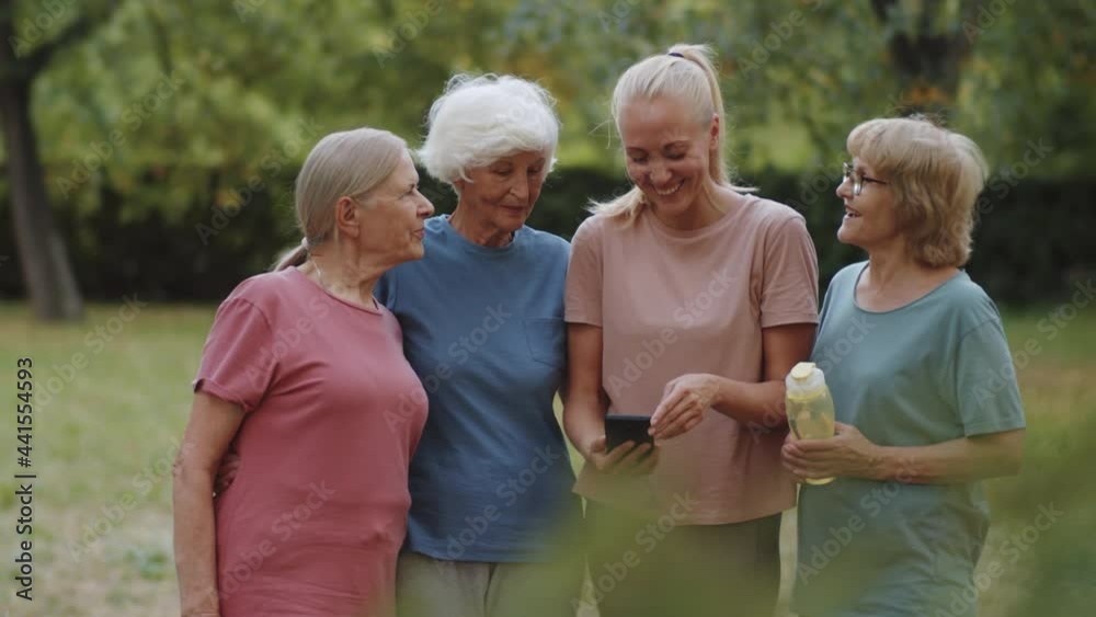 Young female fitness coach smiling and discussing something on smartphone screen with elderly woman in sportswear while standing together in park Young female fitness coach smiling and discussing something on smartphone screen with elderly woman in sportswear while standing together in park