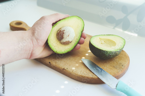 Male caucasian hand holding a half cut haas avocado on a wooden kitchen board with a knife. Photo for step by step avocado salad recipes. The concept of cooking, proper healthy nutrition, vitamins.