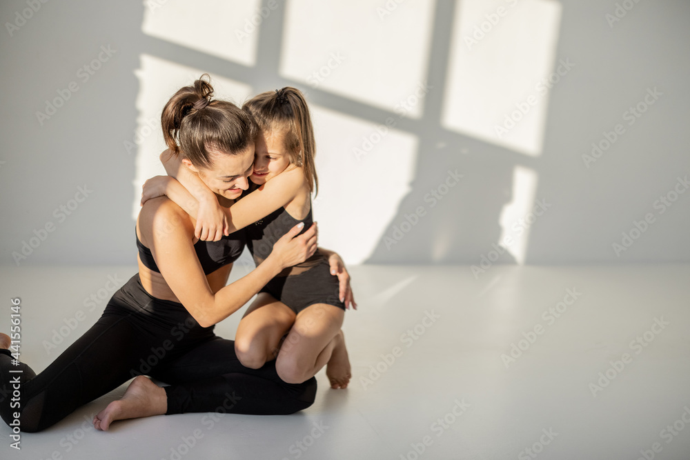 Portrait of a cheerful sports mom and little daughter hug and having fun during sports activities indoors. Close relationships with mother and sports in childhood - obrazy, fototapety, plakaty 