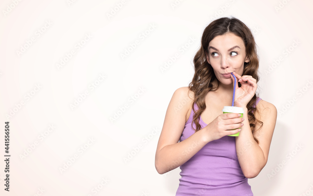 Young happy girl drinking through a straw from a paper cup and looking away, on a light yellow background Young happy girl drinking through a straw from a paper cup and looking away, on a light yellow background