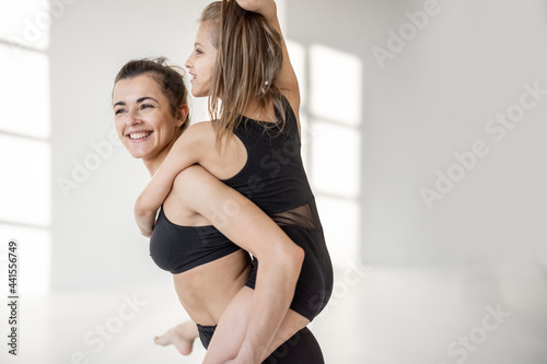 Happy little girl having fun, riding piggyback on her sports mom or trainer during gymnastics training at white classroom