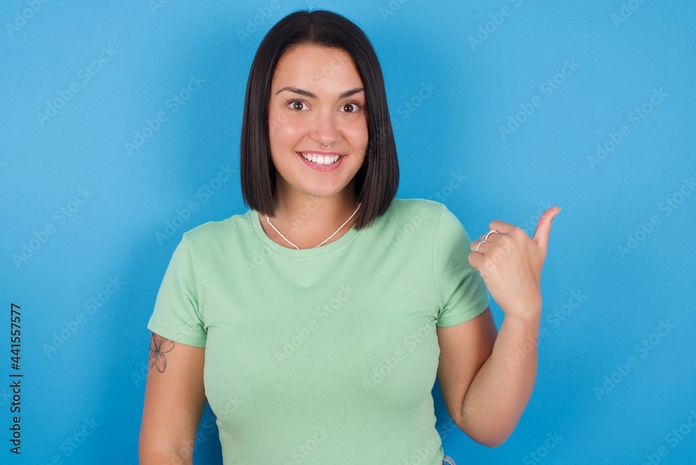 Lovely young beautiful brunette girl with short hair standing against blue background pointing aside with forefinger, showing at copy space having news about bargains Lovely young beautiful brunette girl with short hair standing against blue background pointing aside with forefinger, showing at copy space having news about bargains