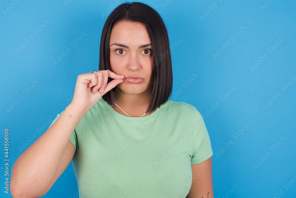young beautiful brunette girl with short hair standing against blue background mouth and lips shut as zip with fingers. Secret and silent, taboo talking. young beautiful brunette girl with short hair standing against blue background mouth and lips shut as zip with fingers. Secret and silent, taboo talking.