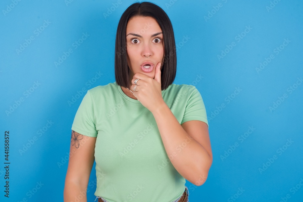 young beautiful brunette girl with short hair standing against blue background Looking fascinated with disbelief, surprise and amazed expression with hands on chin young beautiful brunette girl with short hair standing against blue background Looking fascinated with disbelief, surprise and amazed expression with hands on chin