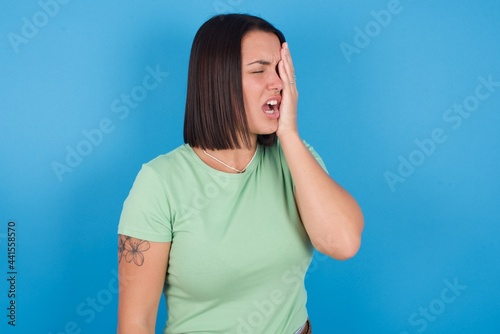 young beautiful brunette girl with short hair standing against blue background Yawning tired covering half face, eye and mouth with hand. Face hurts in pain.