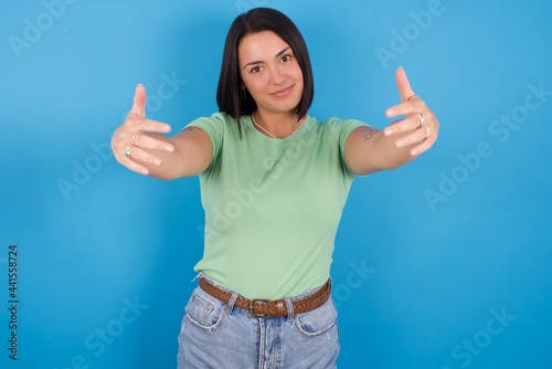 young beautiful brunette girl with short hair standing against blue background looking at the camera smiling with open arms for hug. Cheerful expression embracing happiness.