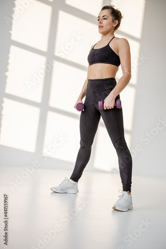 Beautiful and confident athletic woman doing cardio with dumbbells at white room. Training fitness or aerobics alone