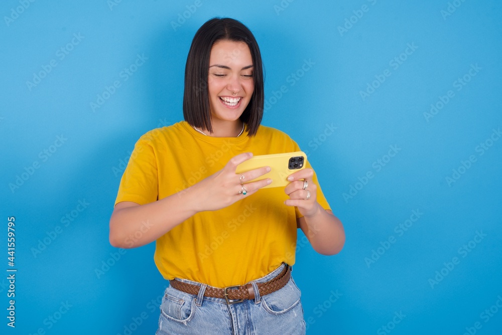 Portrait of an excited young beautiful brunette girl with short hair standing against blue background playing games on mobile phone. Portrait of an excited young beautiful brunette girl with short hair standing against blue background playing games on mobile phone.