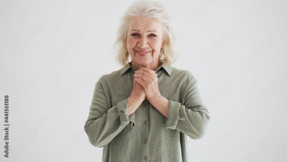 A smiling elder woman is rejoicing while looking to the camera standing isolated over white wall in the studio