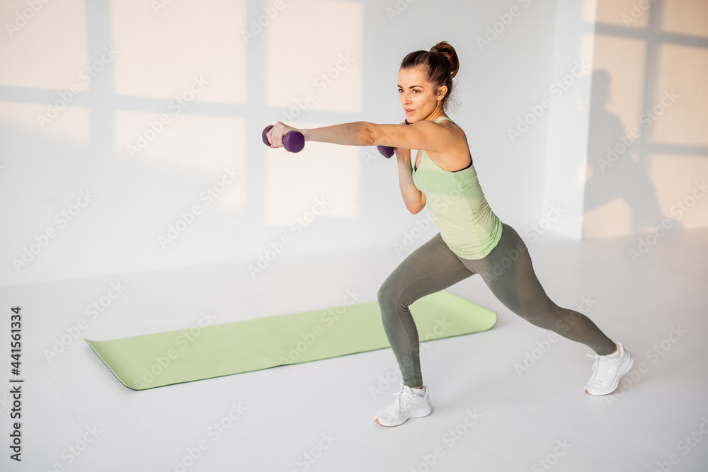 Fototapeta premium Beautiful and confident athletic woman doing cardio with dumbbells at white room. Training fitness or aerobics alone