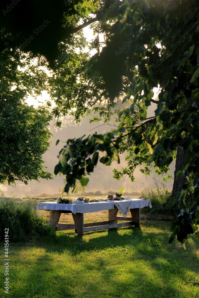 Outdoors summer scene party table on sunset. Old wooden table under trees with food plate. Midsummer celebrating in Latvia, Ligo festive Outdoors summer scene party table on sunset. Old wooden table under trees with food plate. Midsummer celebrating in Latvia, Ligo festive