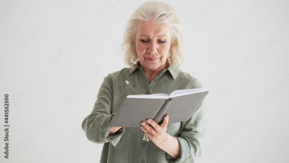 A concentrated elder woman is making notes standing isolated over white wall in the studio A concentrated elder woman is making notes standing isolated over white wall in the studio
