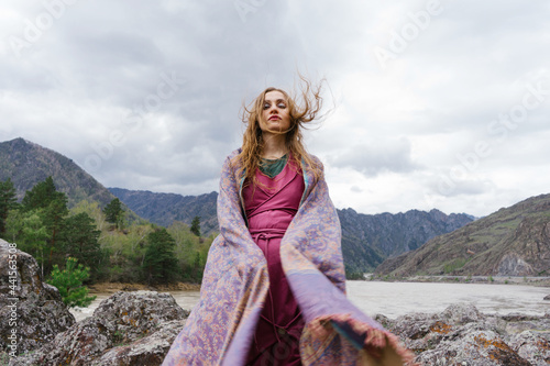 Young beautiful shaman-looking woman standing in the wind in mountains wearing ethnic dress with scarf