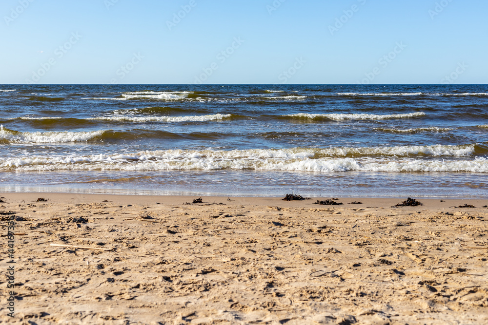 Seashore at Ragakapa Beach in Jurmala, Latvia. Yellow sandy beach where blue Baltic sea waves with white foam are reaching the coast at sunny day. 