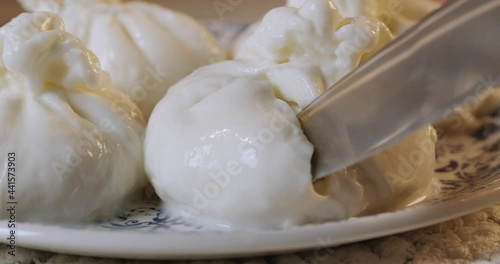 Close up of knife cutting fresh white burrata cheese on plate. Delicate creamy consistency of tasty cheese. Concept of italian cuisine and appetizer.