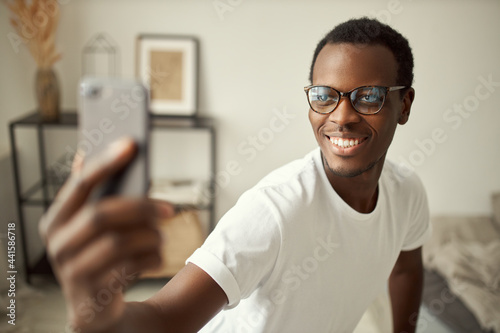 Electronic gadgets concept. Attractive cheerful young dark skinned man wearing white tee and glasses reaching out hand, holding mobile, smiling broadly, taking selfie or having video conference call © Anatoliy Karlyuk