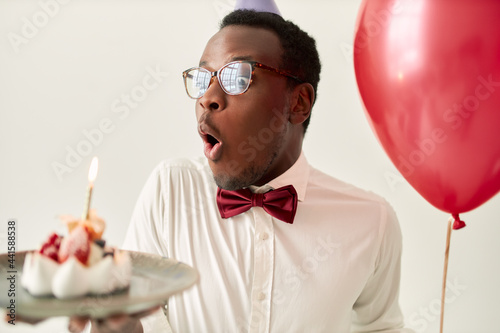 Isolated image of funny fascinated young bearded dark skinned male wearing glasses and bow opening mouth, taking deep breath to blow out burning candle on birthday cake, making wish, holding balloon © Anatoliy Karlyuk