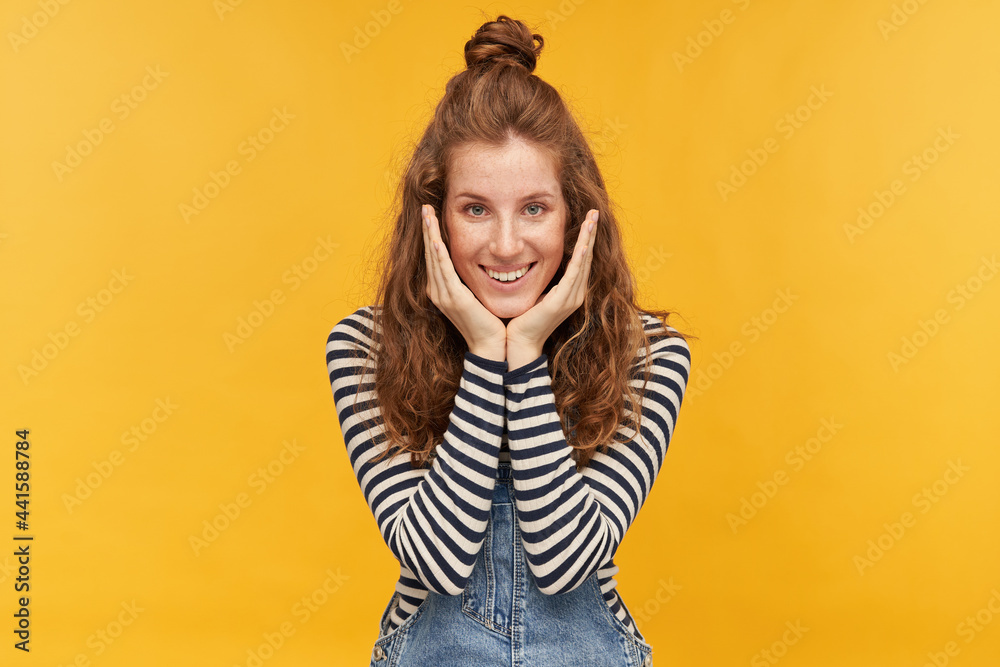young ginger female smiles broadly, keeps her palm together in a pray position. wish herself everything good. wears denim overalls and stripped shirt. isolated over yellow background young ginger female smiles broadly, keeps her palm together in a pray position. wish herself everything good. wears denim overalls and stripped shirt. isolated over yellow background