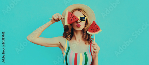 Summer portrait of stylish young woman blowing her lips with lollipop or ice cream shaped slice of watermelon wearing a straw hat on a blue background