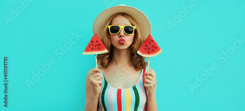 Summer portrait of stylish young woman blowing her lips with lollipop or ice cream shaped slice of watermelon wearing a straw hat on a blue background