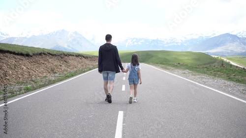 dad and daughter hold hands and walk along the road against the background of mountains