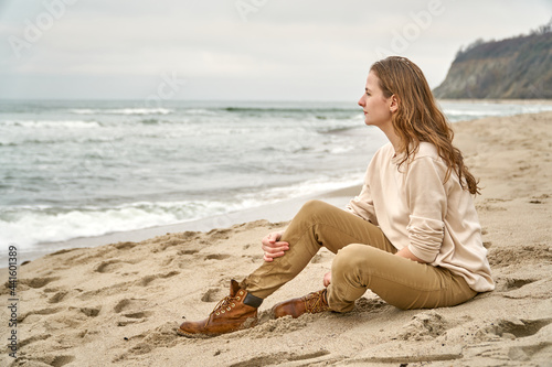 A beautiful woman sits on the sand on the beach and looks into the distance of the sea at the ocean horizon. Fall clothes vacation. Caucasian girl is happy Beige clothes and sand in the fall.