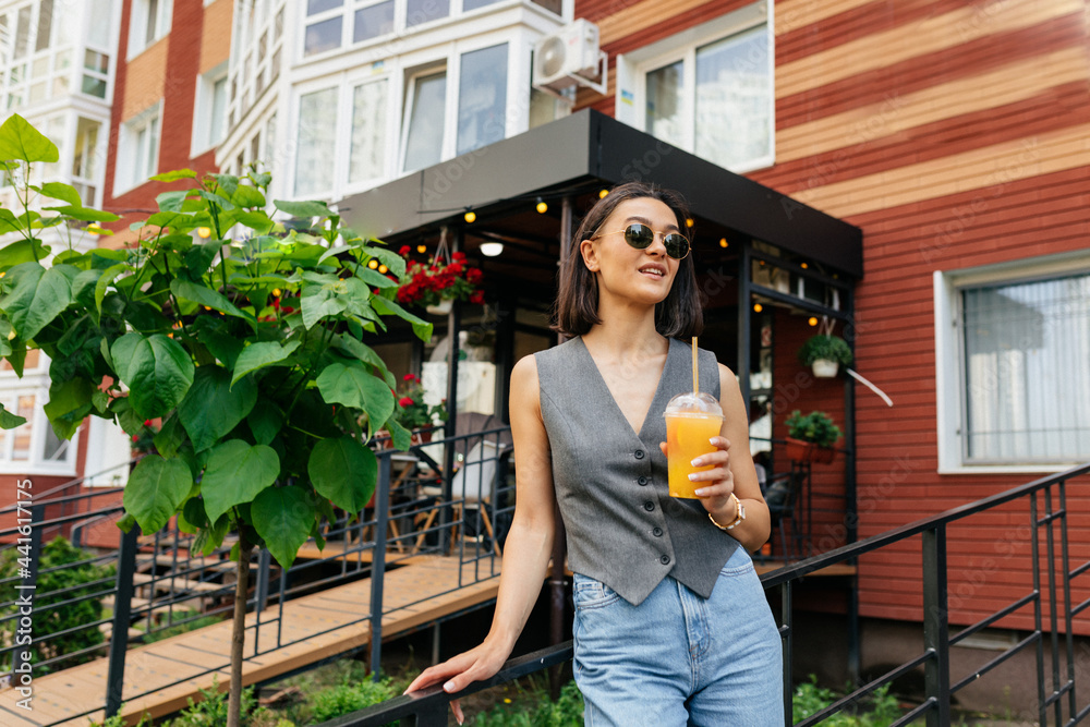 Spectacular stylish lady with orange summer drink looking straight and resting near summer cafe