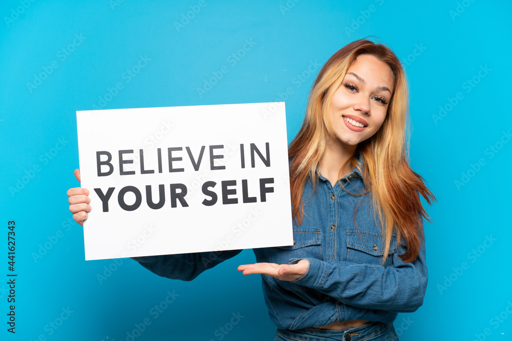 Teenager girl over isolated blue background holding a placard with text Believe In Your Self and pointing it Teenager girl over isolated blue background holding a placard with text Believe In Your Self and pointing it
