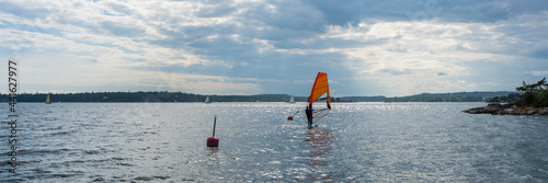 Windsurfing. Beautiful red orange sail. Windsurfer floats on surface of the water in gulf of the Baltic Sea. Dramatic sky with sunbeams. Yachts sailing nearby. Shores of Sweden covered with forest.