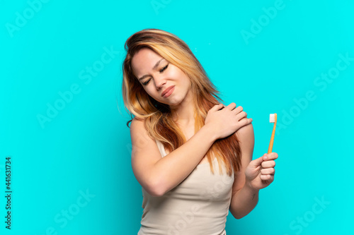 Teenager girl brushing teeth over isolated blue background suffering from pain in shoulder for having made an effort