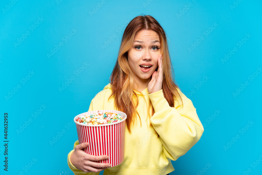 Teenager girl holding popcorns over isolated blue background with surprise and shocked facial expression