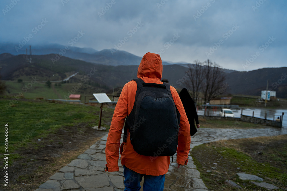 Naklejka premium A person in an orange raincoat with a backpack walks in the rain in the mountains