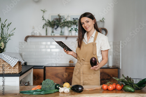 beautiful young brunette woman prepares in the kitchen according to a recipe from a notebook a healthy meal from fresh organic vegetables looks at the camera