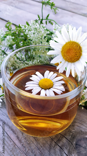 Chamomile tea in a glass cup close-up on a wooden background. Summer still life with wildflowers and medicinal chamomile drink. Floral vertical background.