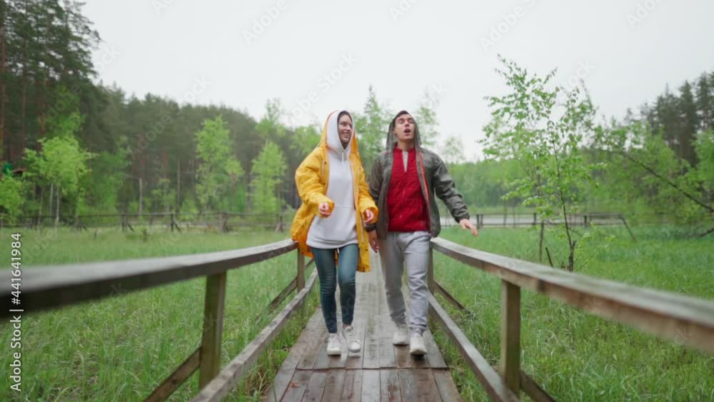 Happy young couple putting on hoods of raincoats while walking down wooden path in countryside and talking in rain. Cheerful friends enjoying travel despite of weather, slow motion shot Happy young couple putting on hoods of raincoats while walking down wooden path in countryside and talking in rain. Cheerful friends enjoying travel despite of weather, slow motion shot