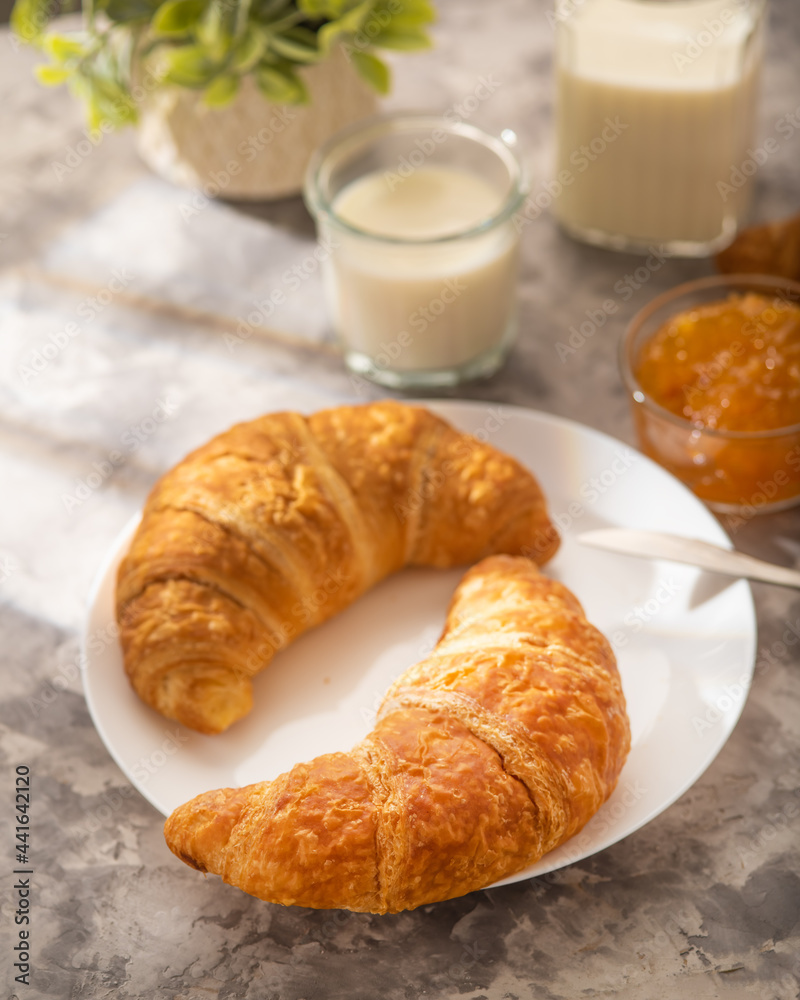 Croissants on a white earthenware plate. In the background, orange jam, milk in a glass jug and glass. White background. High angle view. There are no people in the photo. Croissants on a white earthenware plate. In the background, orange jam, milk in a glass jug and glass. White background. High angle view. There are no people in the photo.
