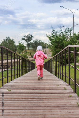 A little child is running away on wooden pathway with a fence outdoors in the park © Anna