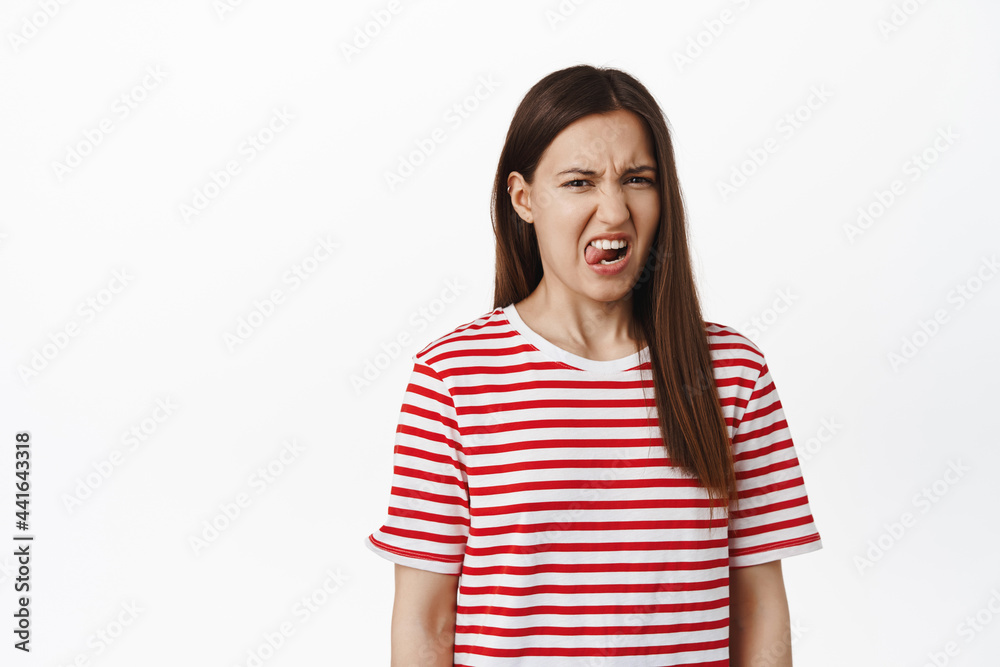 Disgusting. Young 20s girl student express dislike or aversion, show tongue, grimacing and squinting from bad taste, standing displeased against white background Disgusting. Young 20s girl student express dislike or aversion, show tongue, grimacing and squinting from bad taste, standing displeased against white background