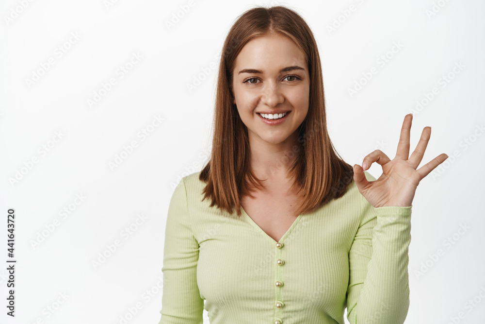 Okay. Smiling blond girl shows ok zero gesture, no problem, yes or alright, approve smth, give positive feedback, like and agree, recommending place, white background Okay. Smiling blond girl shows ok zero gesture, no problem, yes or alright, approve smth, give positive feedback, like and agree, recommending place, white background