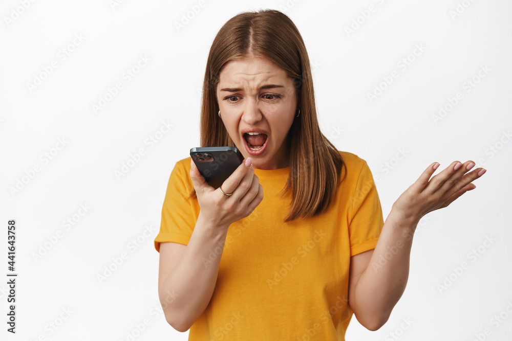 Angry girl shouting at smartphone voice message, talk on speakerphone and screaming at mobile phone dynamic, shaking hands frustrated, standing against white background Angry girl shouting at smartphone voice message, talk on speakerphone and screaming at mobile phone dynamic, shaking hands frustrated, standing against white background