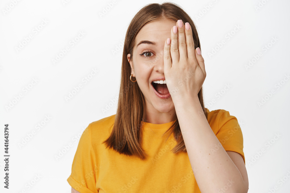Portrait of happy young woman cover half of face, laughing and smiling amazed at camera, stare surprised, standing in yellow t-shirt against white background Portrait of happy young woman cover half of face, laughing and smiling amazed at camera, stare surprised, standing in yellow t-shirt against white background