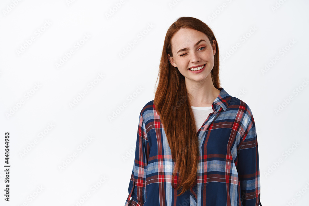 Positive caucasian woman with red long hair, freckles and clean skin, smiling and winking at camera happy, standing in plaid shirt against white background Positive caucasian woman with red long hair, freckles and clean skin, smiling and winking at camera happy, standing in plaid shirt against white background