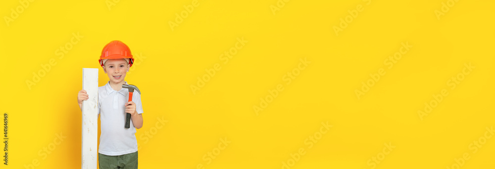 Smiling child with a construction tool (hammer) and building materials stands on a yellow background, he is happy. Free space for an inscription, a banner Smiling child with a construction tool (hammer) and building materials stands on a yellow background, he is happy. Free space for an inscription, a banner