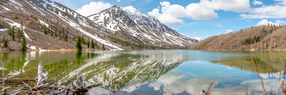 Panoramic view of St Elias Lake in northern Canada with calm, reflective water below snow capped peaks. 
